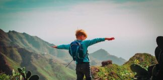 Child with backpack stands on mountain arms outstretched