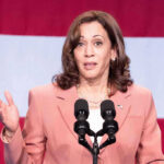 Trump YANKS Harris’ Protection—Chaos Erupts Woman speaks at podium with red and white background.