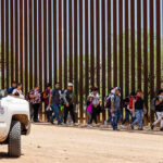 Late-Night Judge BLOCKS Trump’s CHILD Deportation Move People walking beside tall fence and border patrol vehicle.