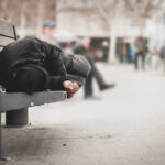 NEW Blood-Sharing DRUG Rampant—HIV Spikes! Person sleeping on a bench in a public area.