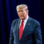 Trump in HOSPITAL – Questions Raised! Man in suit with red tie speaking on stage.