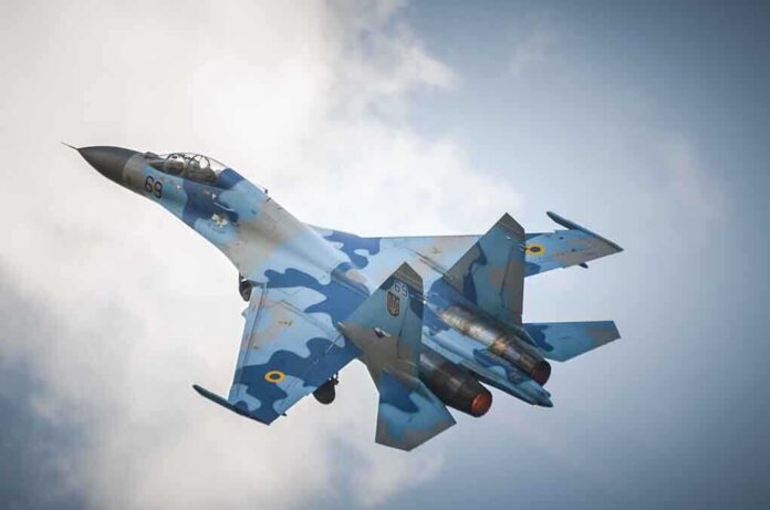A blue camouflage fighter jet performing an aerial maneuver against a cloudy sky