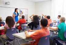 RACIST Teacher’s SHOCKING Classroom Stunt Sparks Outrage! Teacher in a blue dress instructing students in a classroom with hands raised