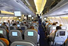 Midair Mayhem – Maniac STRANGLES Flight Attendant! Interior view of an airplane with passengers seated and using in-flight entertainment screens