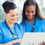 Two nurses in blue scrubs smiling while looking at a tablet