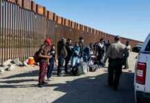 Group of people standing by a border fence with officers and vehicles
