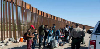 Group of people standing by a border fence with officers and vehicles