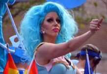 Dem Candidate’s Drag Queen OBSESSION Gets Weirder Drag performer with bright blue wig giving thumbs up at parade