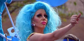 Drag performer with bright blue wig giving thumbs up at parade