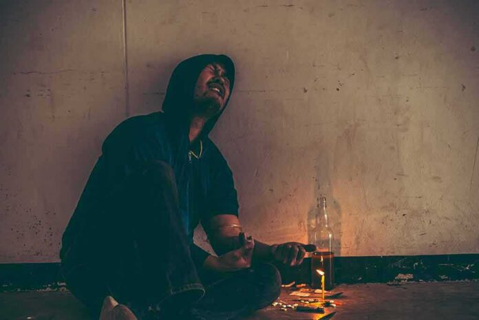Person in hoodie sitting on floor with drug paraphernalia