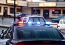 Dem Candidate ARRESTED – Handcuffed in Police Car Police car with flashing red and blue lights in a parking lot