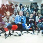 Row of microphones at a press conference with audience and cameras behind
