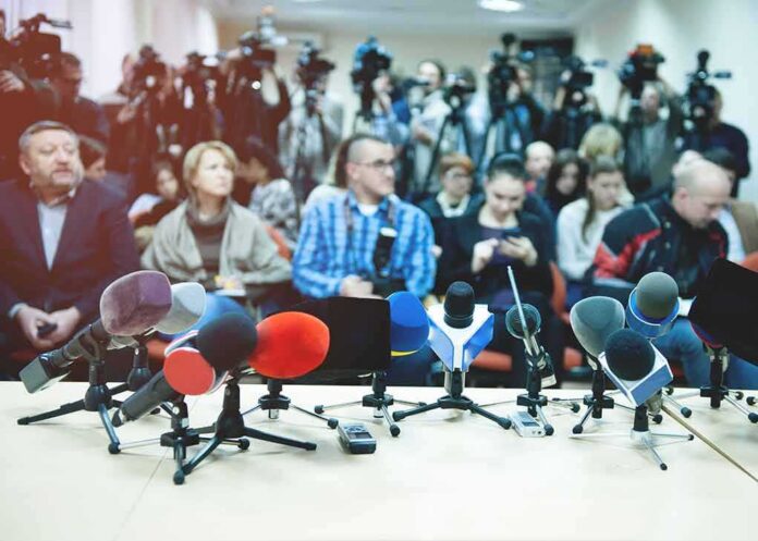 Row of microphones at a press conference with audience and cameras behind