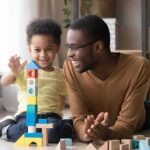 Father and child building a tall tower with colorful wooden blocks on the floor