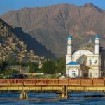 White mosque with blue domes by a river and bridge, backed by rugged mountains