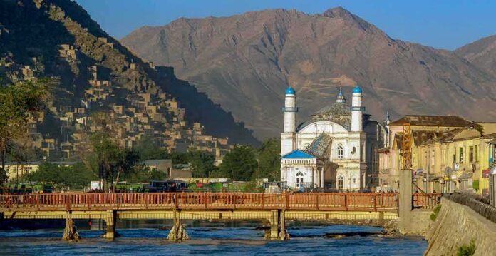 White mosque with blue domes by a river and bridge, backed by rugged mountains