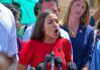 Woman in red dress speaking passionately at outdoor press conference
