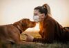 Woman in mask touching noses with brown dog at sunset