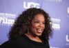 Oprah Tells Audience To CELEBRATE Child Killer Mothers smiling woman with curly hair on a blue step-and-repeat backdrop