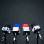 Four broadcast microphones on stands against a dark background