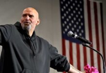 Fetterman Swears GOP Allegiance – Rattles Dems Bald man speaking at a microphone with a U.S. flag in the background