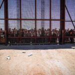 Crowd stands behind tall metal fence at border crossing