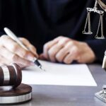 Podcaster Indicted Over ‘Kill Trump’ Rants Judge writing at desk with gavel and Lady Justice statue