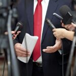 Man in suit with red tie speaking to reporters holding microphones