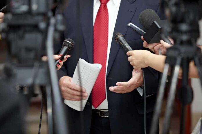 Man in suit with red tie speaking to reporters holding microphones